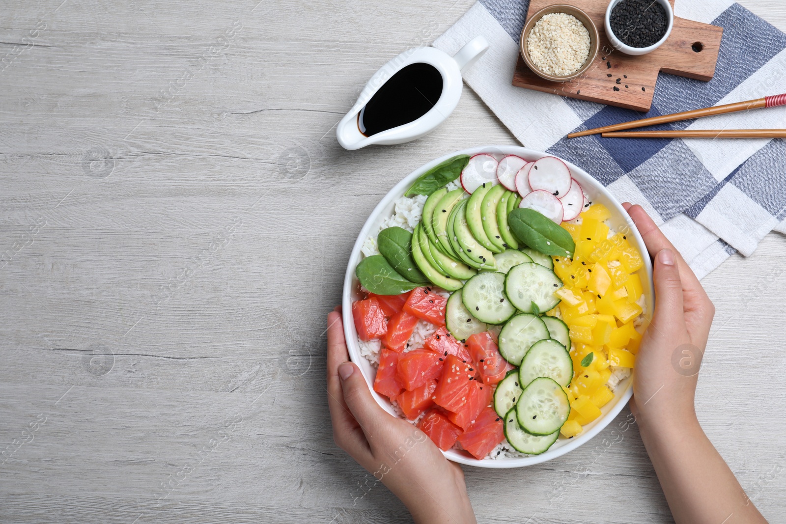 Photo of Woman holding delicious poke bowl with salmon and vegetables at wooden table, top view. Space for text