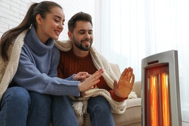 Photo of Young couple warming hands near electric heater at home