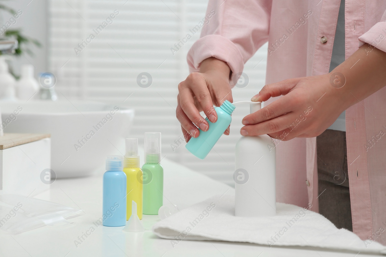 Photo of Woman pouring cosmetic product into plastic bottle at white countertop in bathroom, closeup. Bath accessories