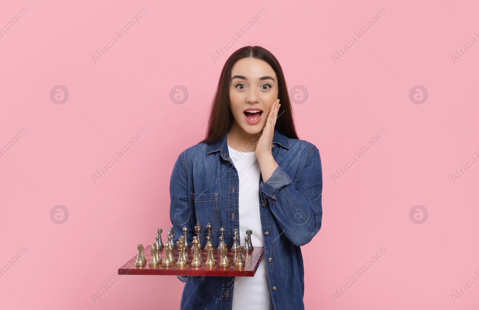 Photo of Emotional woman holding chessboard with game pieces on pink background