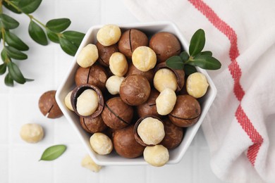 Tasty Macadamia nuts in bowl and green twigs on white table, flat lay