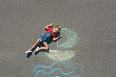 Photo of Cute little child lying near chalk drawing of boat on asphalt, top view