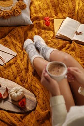 Photo of Woman in warm socks with cup of hot drink resting on knitted blanket, closeup