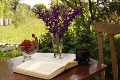 Photo of Open book, cup of tea, strawberries and beautiful wildflowers on table in garden