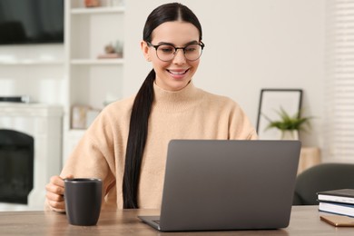 Photo of Happy woman with cup of drink working on laptop at wooden desk in room