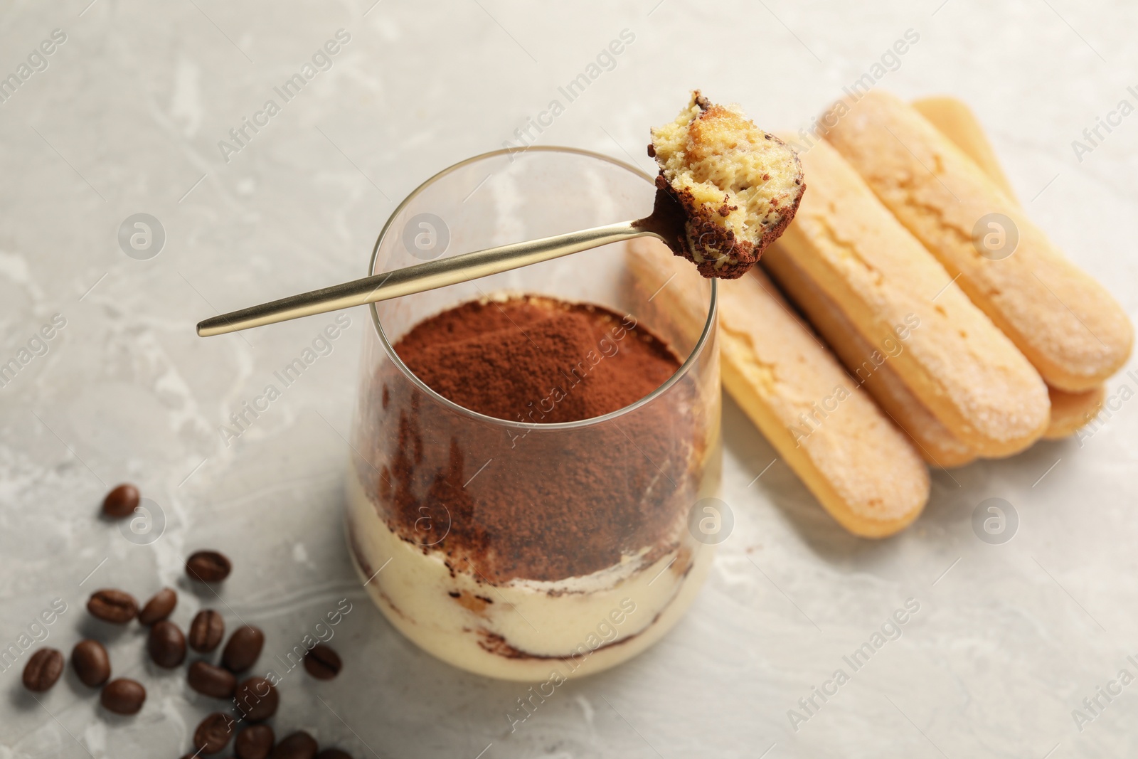 Photo of Tasty tiramisu in glass, coffee beans, spoon and biscuits on light grey table, closeup