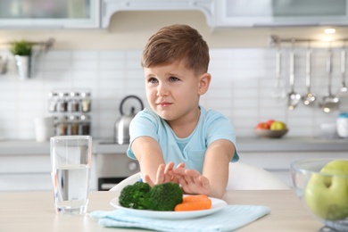 Adorable little boy refusing to eat vegetables at table in kitchen