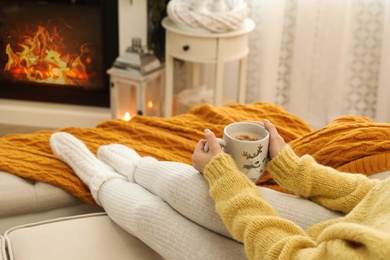 Woman with cup of hot drink resting on sofa near fireplace in living room, closeup