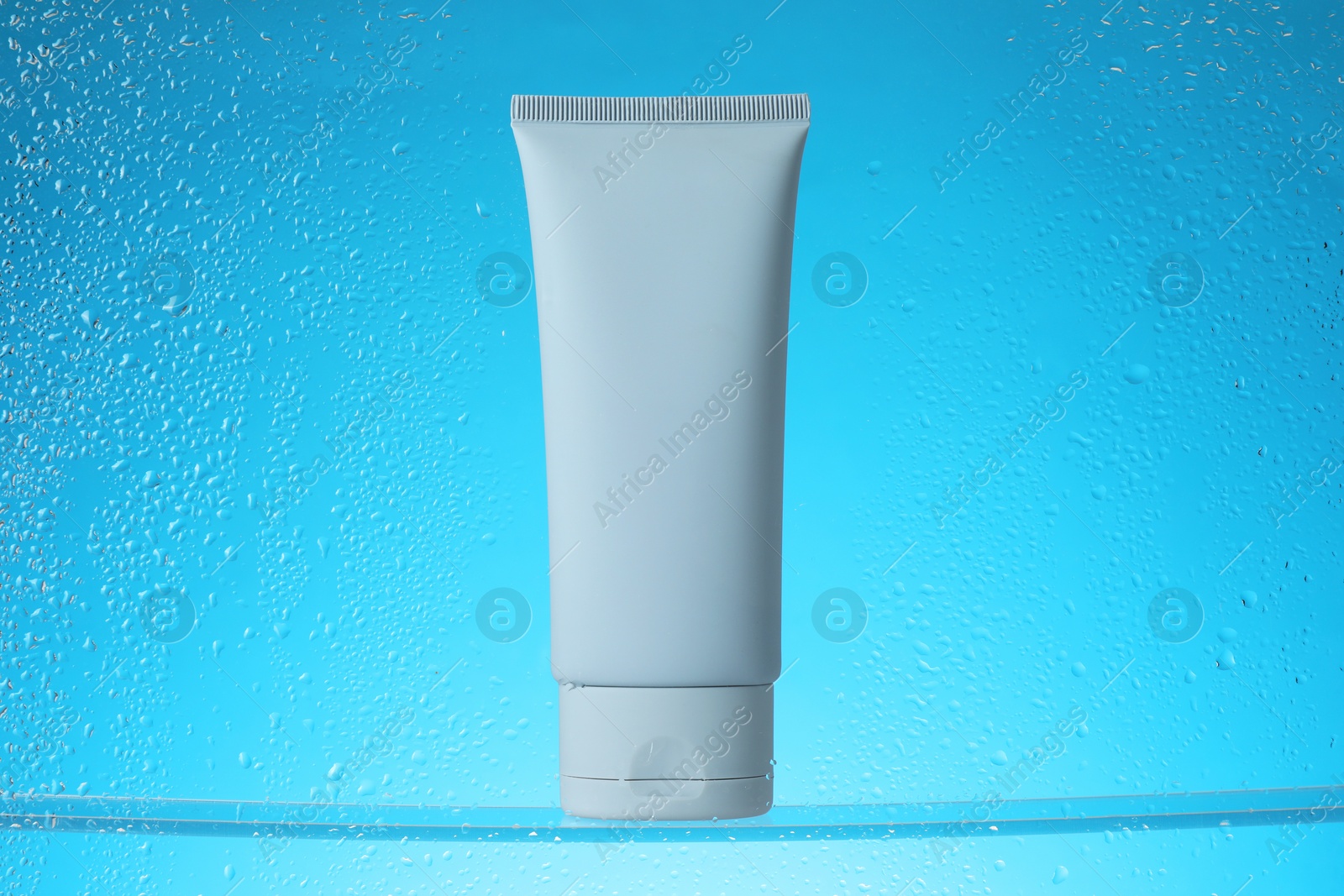 Photo of Tube with moisturizing cream on light blue background, view through wet glass