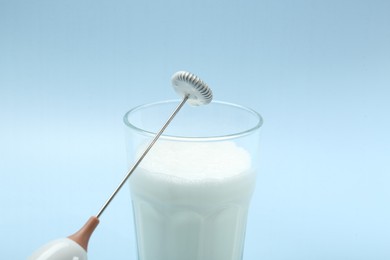 Mini mixer (milk frother) and whipped milk in glass on light blue background, closeup