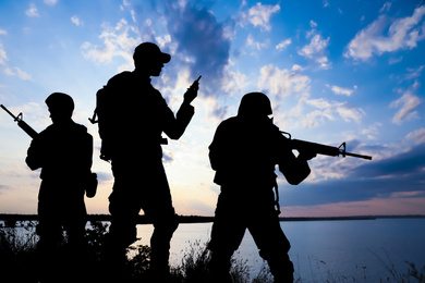 Image of Silhouettes of soldiers with assault rifles and portable radio transmitter patrolling outdoors. Military service