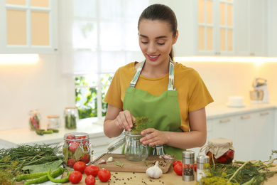 Woman putting dill into pickling jar at table in kitchen