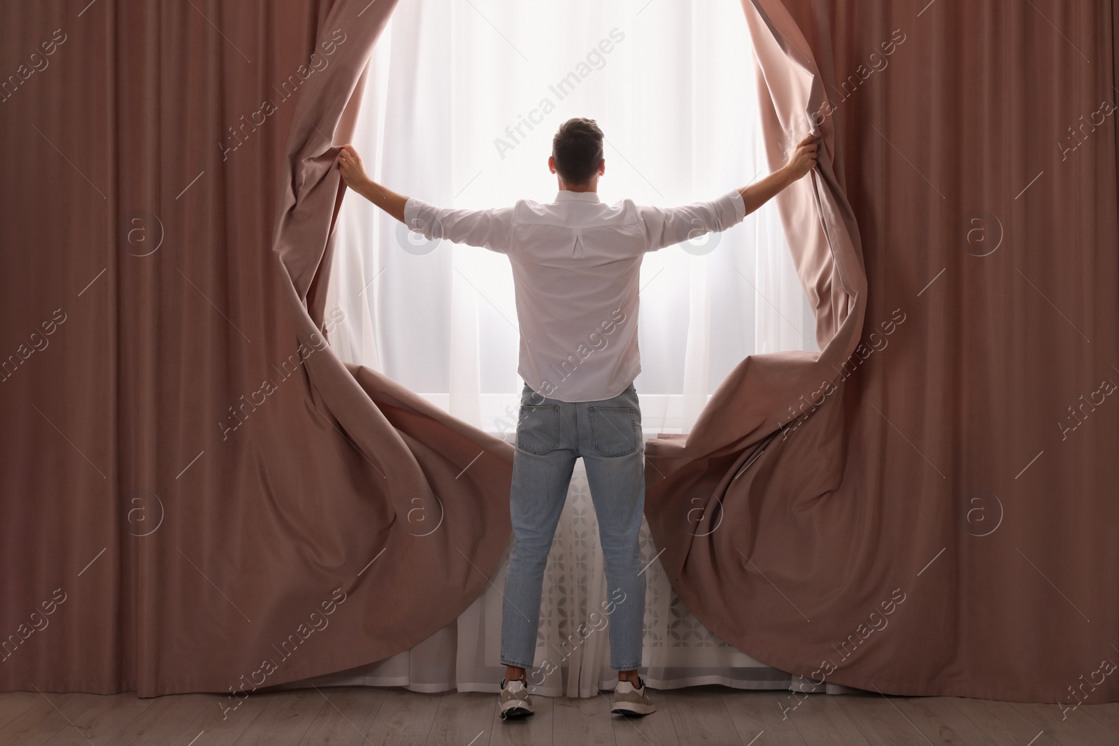 Photo of Man opening window curtains at home, back view