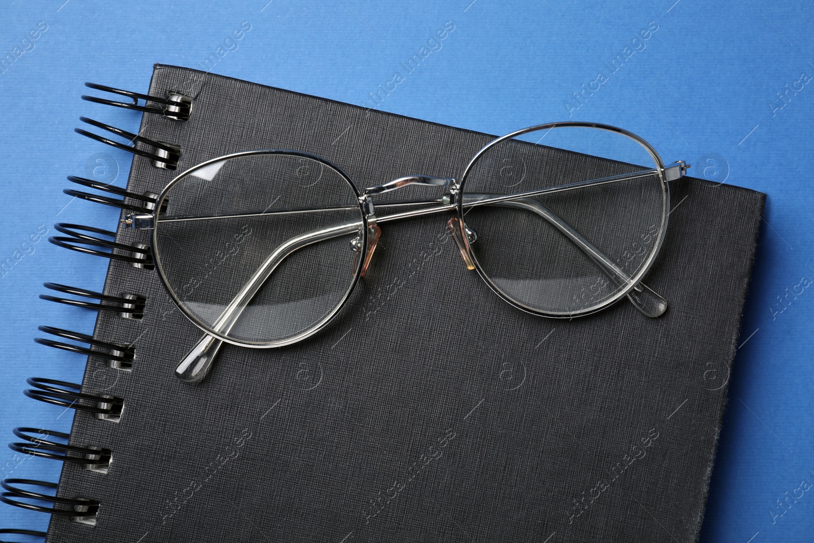Photo of Stylish pair of glasses with metal frame and notebook on blue background, top view