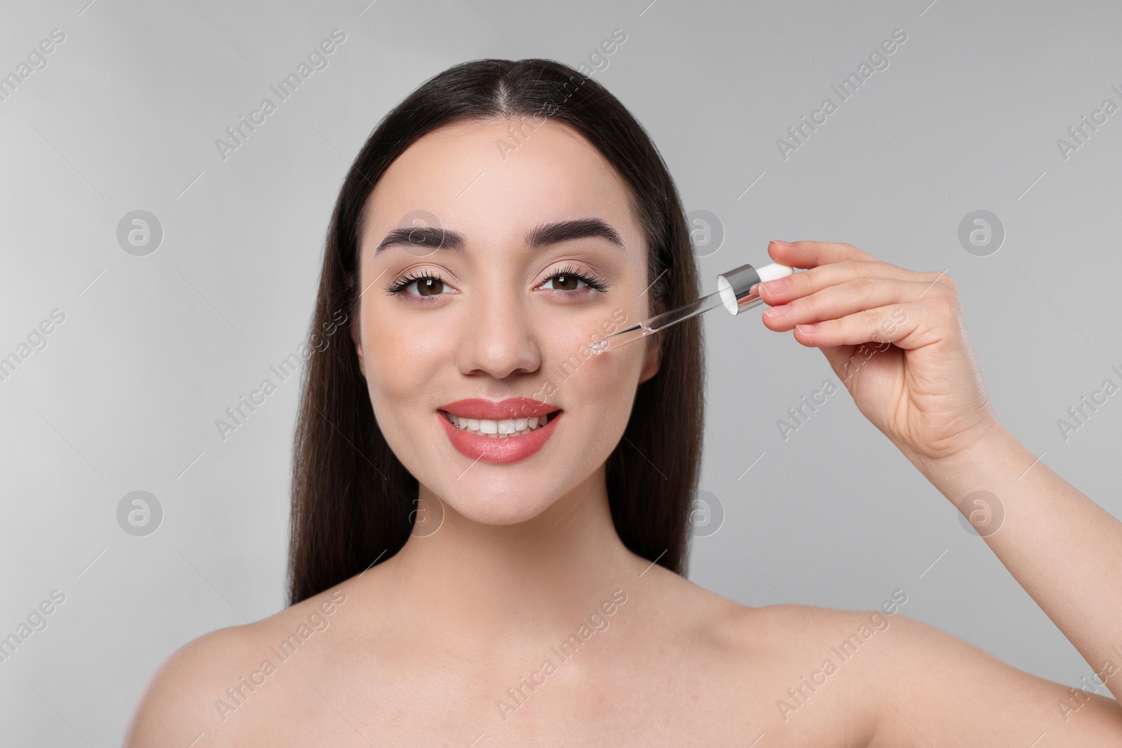 Photo of Happy young woman applying essential oil onto face on light grey background
