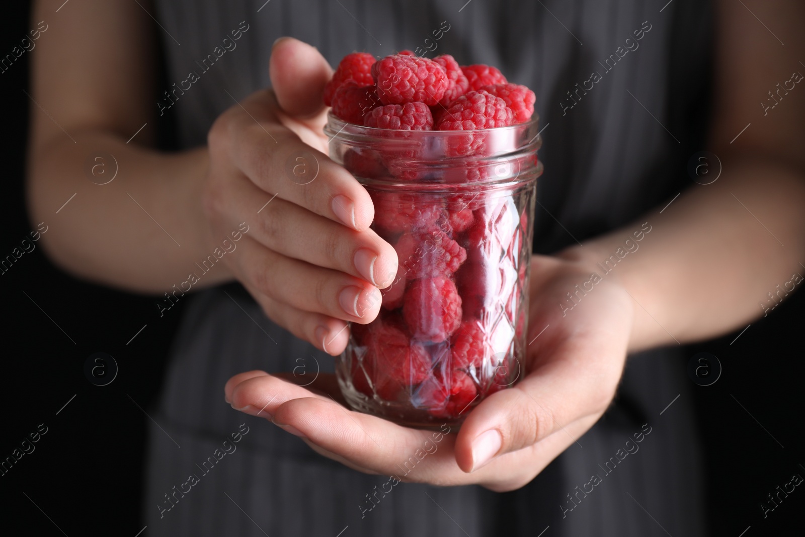 Photo of Woman holding glass jar of delicious ripe raspberries on black background, closeup