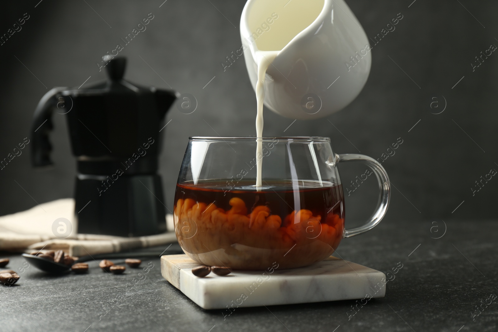 Photo of Pouring milk from pitcher into glass cup with coffee at dark textured table, closeup