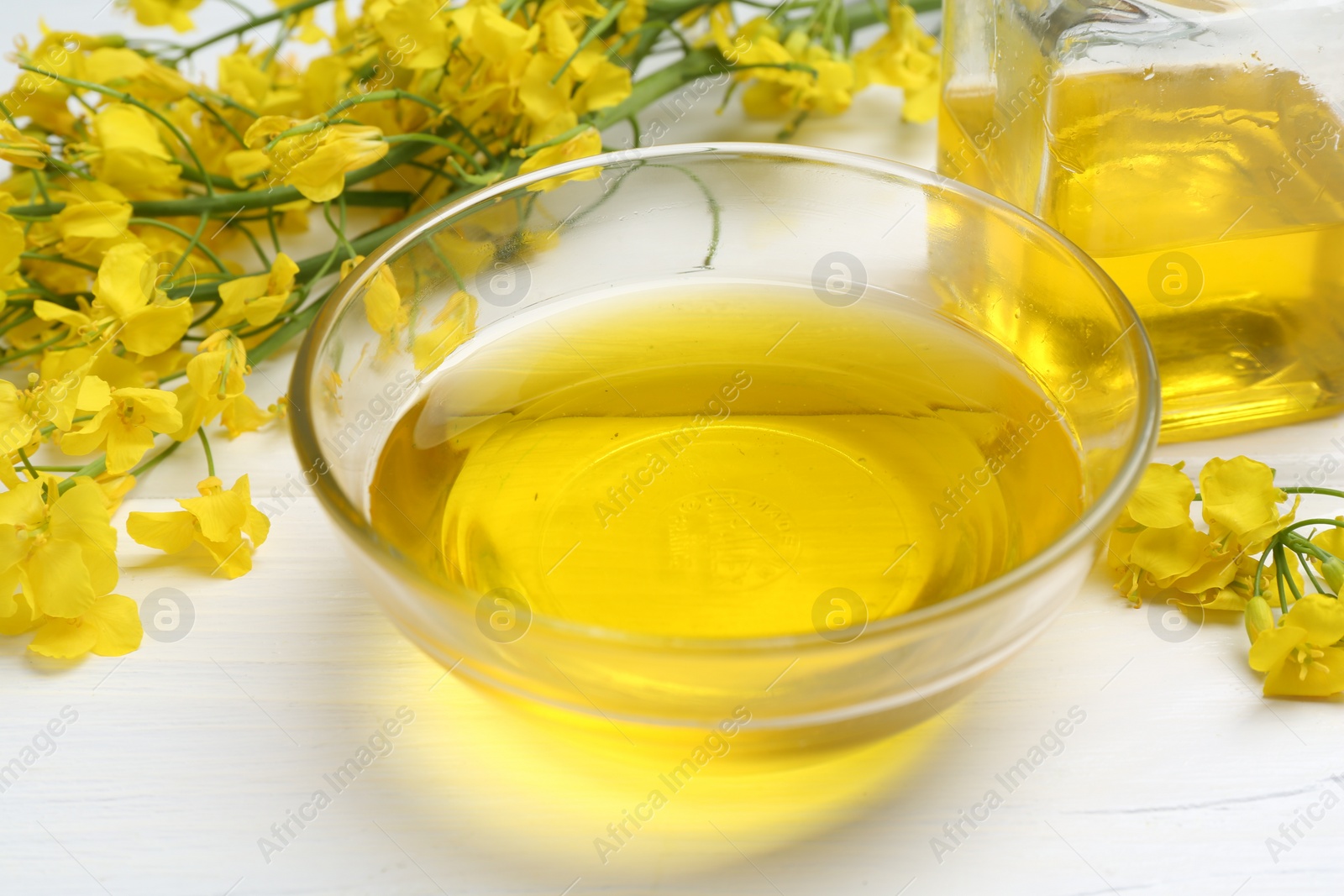 Photo of Rapeseed oil in bowl and beautiful yellow flowers on white wooden table, closeup