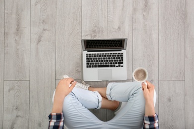 Photo of Young man with cup of coffee using laptop while sitting on floor, top view