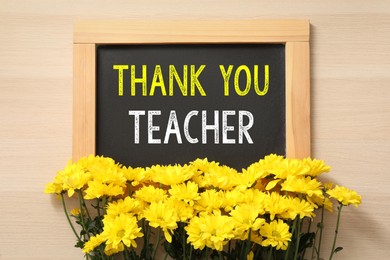 Blackboard with phrase Thank You Teacher and flowers on wooden table, flat lay