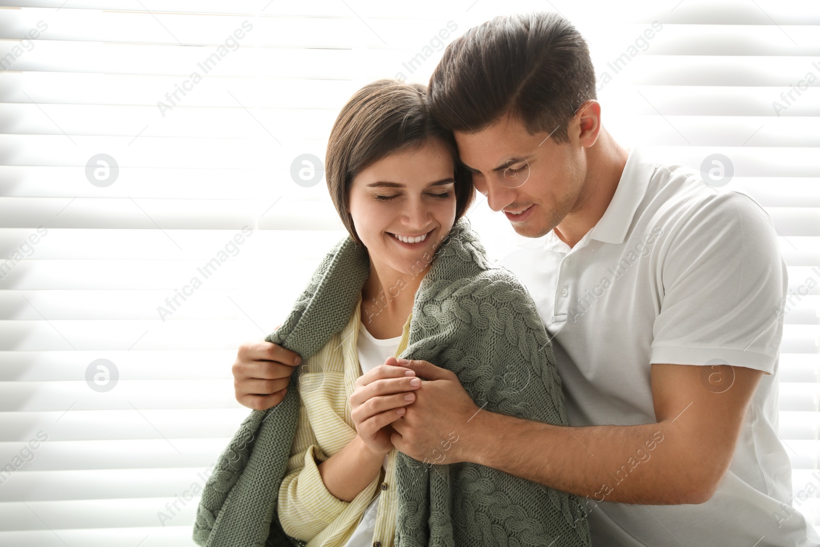 Photo of Man covering his woman with warm green plaid near window indoors