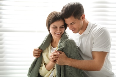 Man covering his woman with warm green plaid near window indoors