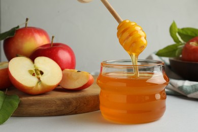 Dripping sweet honey from dipper into jar and fresh apples on white table