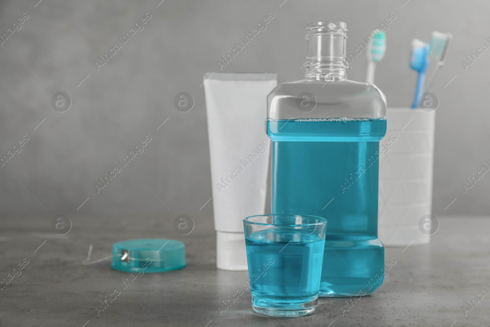 Photo of Bottle and glass with mouthwash near other oral hygiene products on grey table, space for text