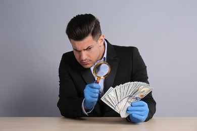 Photo of Expert authenticating 100 dollar banknotes with magnifying glass at table on light grey background. Fake money concept