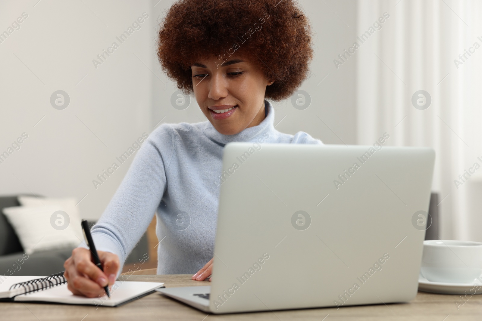 Photo of Beautiful young woman using laptop and writing in notebook at wooden desk in room