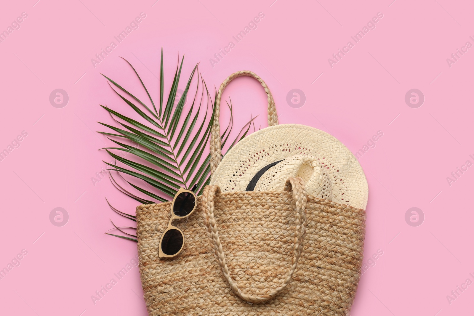 Photo of Elegant woman's straw bag with hat, tropical leaf and sunglasses on pink background, top view