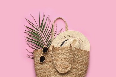 Elegant woman's straw bag with hat, tropical leaf and sunglasses on pink background, top view