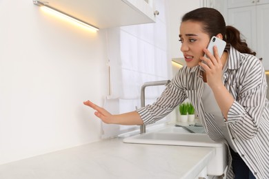 Photo of Confused woman talking on phone and looking at wall in kitchen