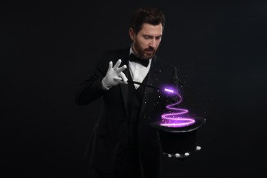 Image of Magician showing trick with wand and top hat on dark background. Fantastic light coming out of hat