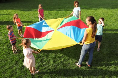 Photo of Group of children and teachers playing with rainbow playground parachute on green grass. Summer camp activity