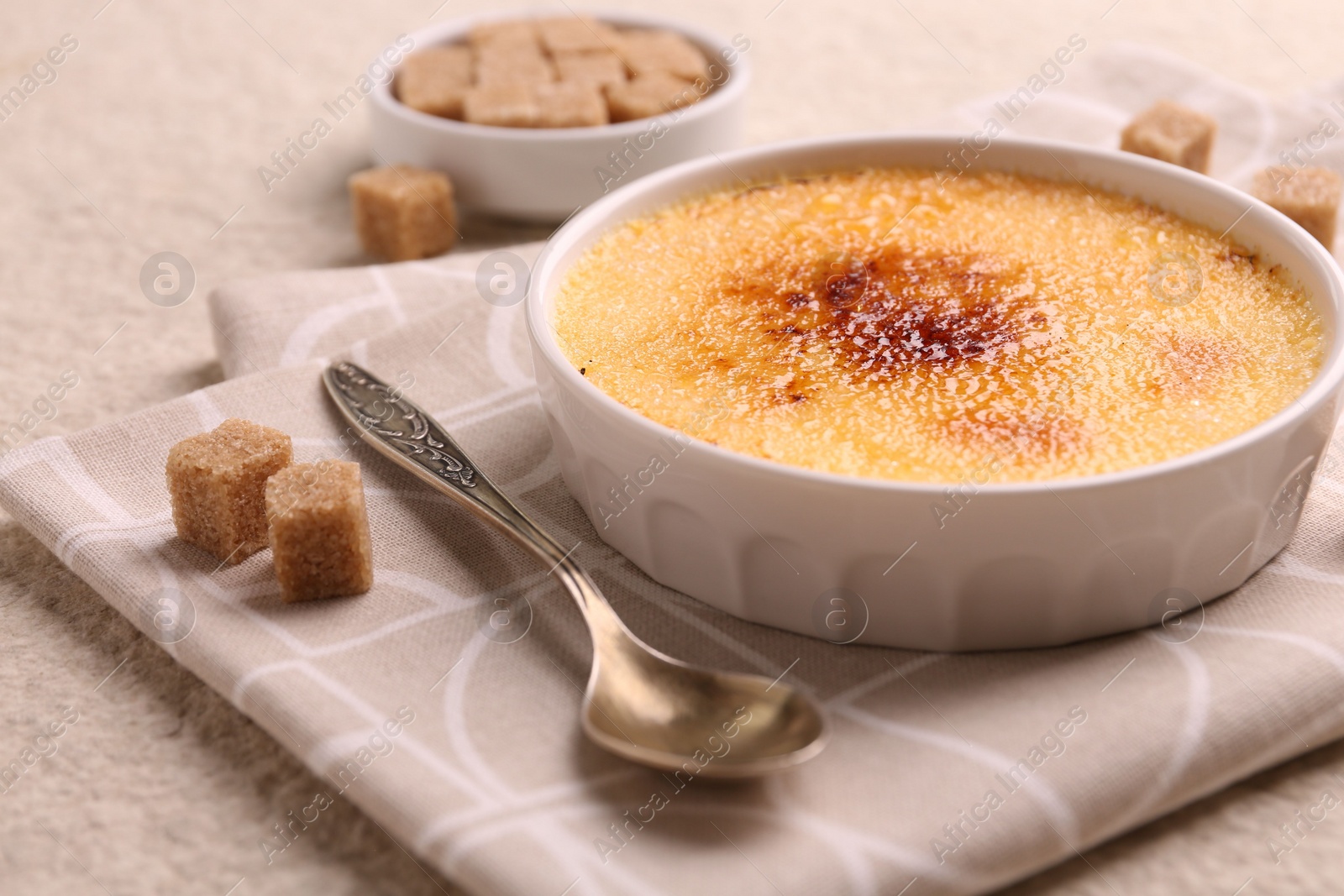 Photo of Delicious creme brulee in bowl, sugar cubes and spoon on textured table, closeup