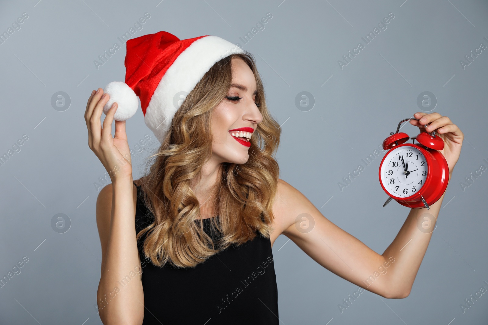 Photo of Happy young woman wearing Santa hat with alarm clock on grey background. Christmas time