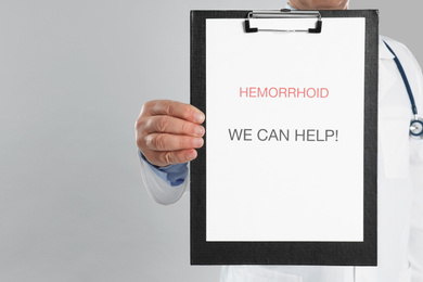 Photo of Doctor holding clipboard with words HEMORRHOID WE CAN HELP on light grey background, closeup