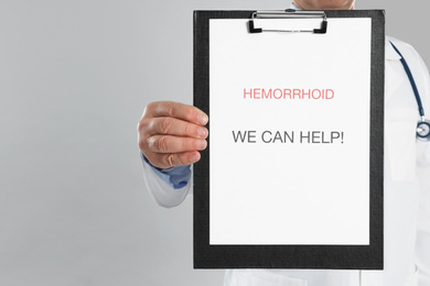 Doctor holding clipboard with words HEMORRHOID WE CAN HELP on light grey background, closeup