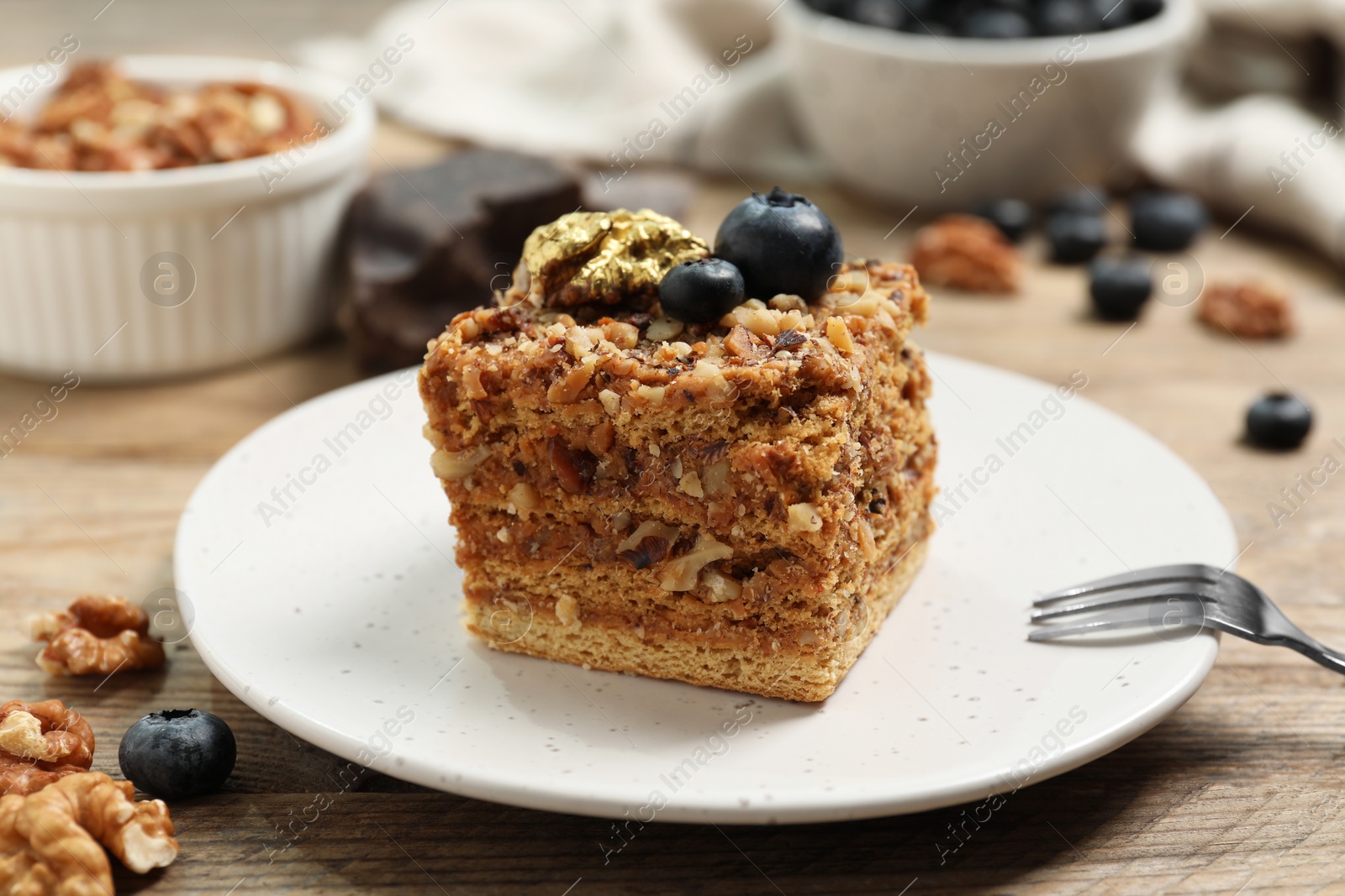 Photo of Piece of delicious layered honey cake with blueberries and nuts served on wooden table, closeup