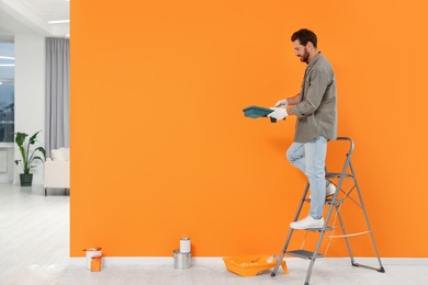 Photo of Designer with container and painting equipment near freshly painted orange wall indoors, space for text