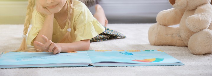 Image of Cute little girl lying on cozy carpet with book and teddy bear at home, closeup. Banner design
