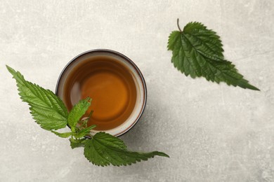Cup of aromatic nettle tea and fresh green leaves on light grey table, flat lay