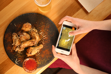 Photo of Woman playing game using smartphone at table with tasty BBQ wings, top view
