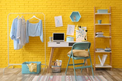 Photo of Fashion designer's workplace with wooden furniture and laptop near yellow brick wall. Stylish interior