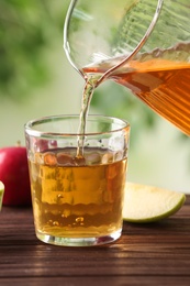 Pouring apple juice into glass on wooden table against blurred green background