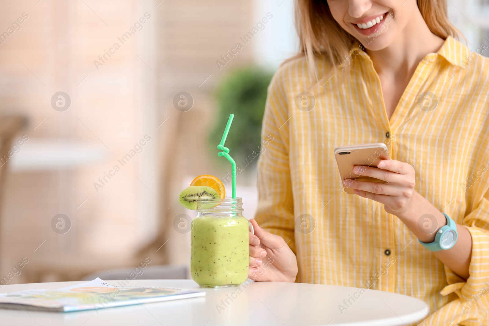 Photo of Young woman using mobile phone while drinking tasty healthy smoothie at table, indoors