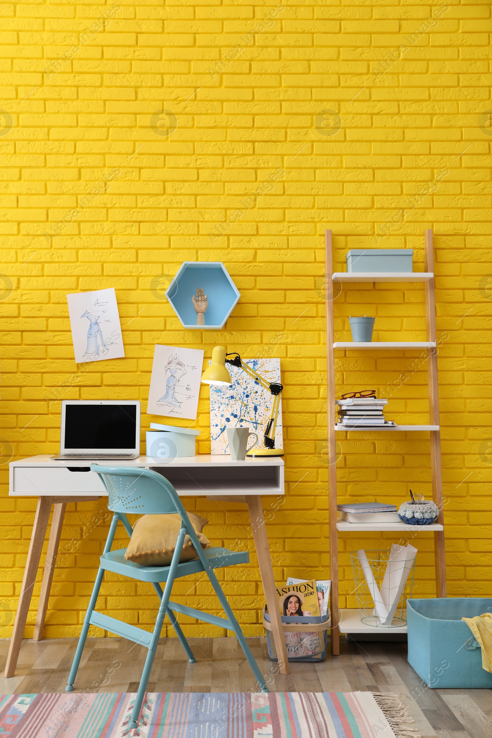 Photo of Fashion designer's workplace with wooden furniture and laptop near yellow brick wall. Stylish interior