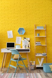 Photo of Fashion designer's workplace with wooden furniture and laptop near yellow brick wall. Stylish interior