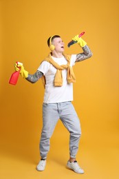 Photo of Handsome young man with brush and bottle of detergent singing on orange background